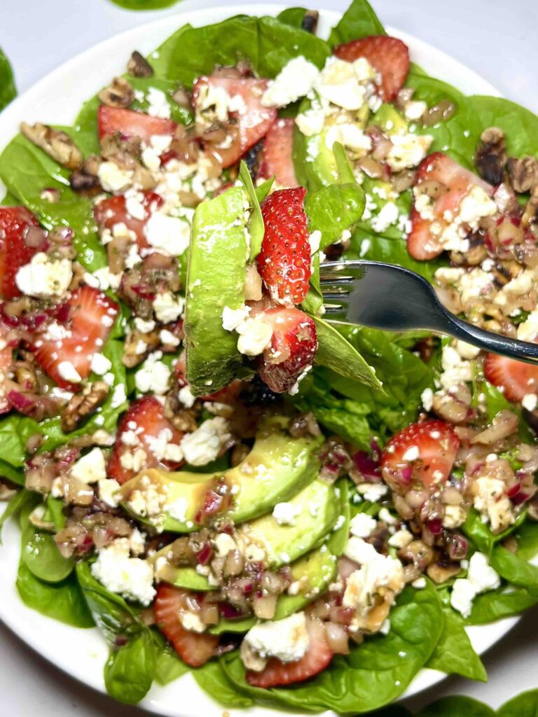 close-up of fork picking up bite of spinach strawberry salad with plate of salad in background