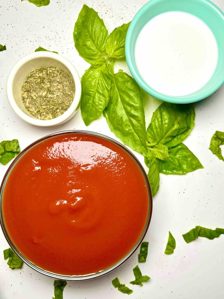 cup of tomato sauce next to cup of coconut cream and cup of seasonings, with basil leaves scattered between, seen from above